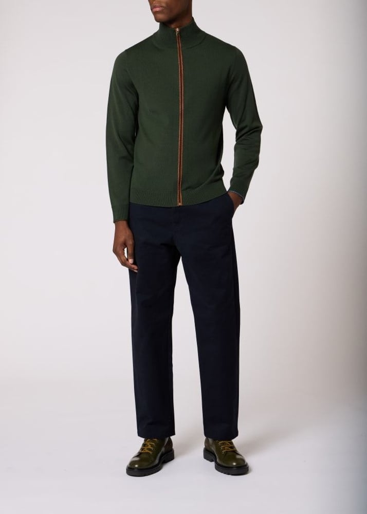 A model wearing a dark green long-sleeve zip-up mock neck sweater with a contrasting gold zipper, paired with navy trousers and dark green leather shoes, against a plain light gray background.